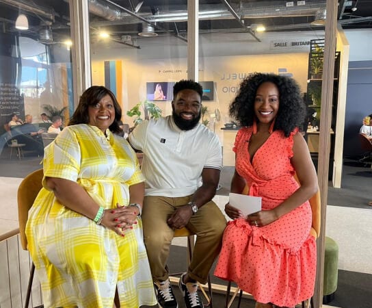Three individuals seated together, wearing brightly colored outfits and smiling during an on-camera discussion.