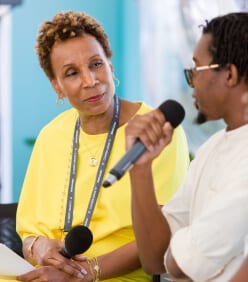 A woman in a bright yellow dress looks on as a man with a microphone speaks at an event.
