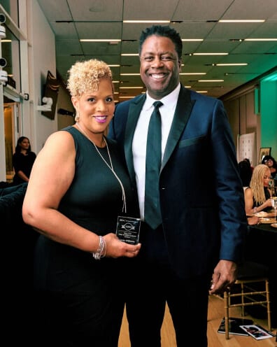 Two individuals in formal black attire pose for a photo; the woman holds an award plaque.