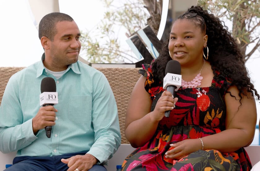 A man in a light green shirt and a woman in a floral black and red dress, both holding microphones and engaged in a conversation during an event.