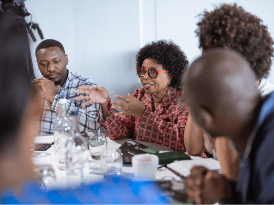 A woman with short curly hair and red-framed glasses leads a lively roundtable discussion with several people seated around her.