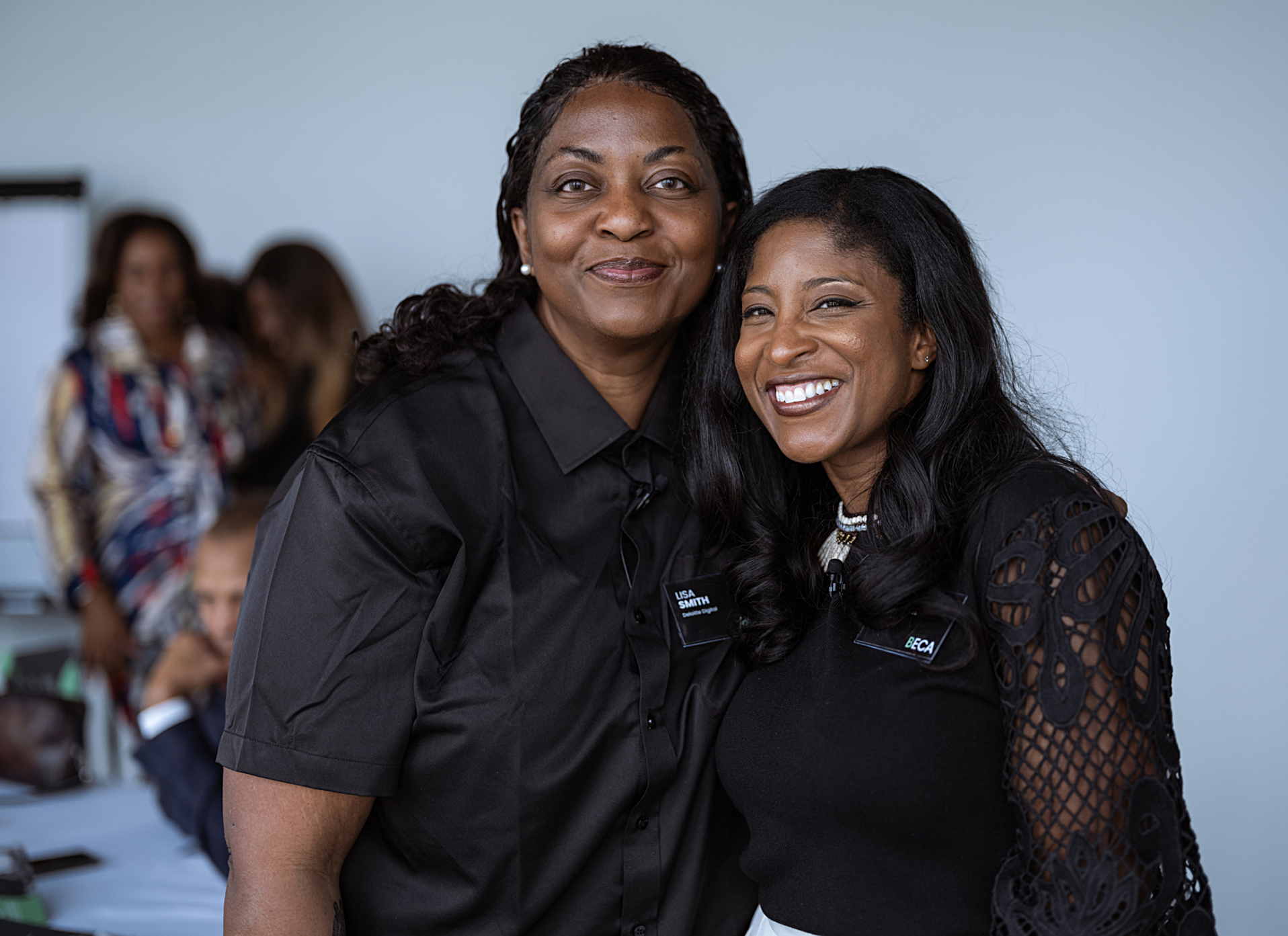 Two women in matching black tops and name badges smile side by side in a bright meeting space, while others gather in the blurred background.