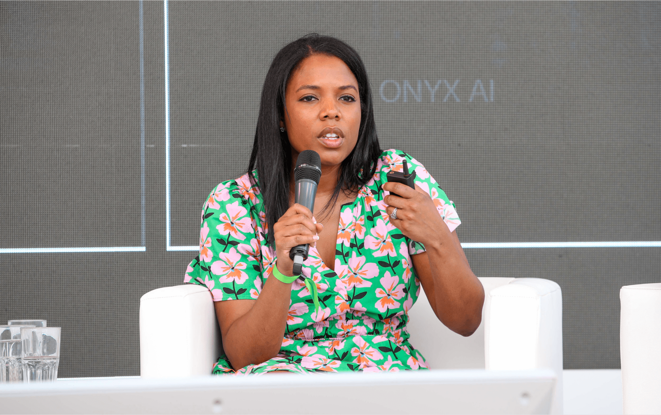 A woman in a floral green dress holding a microphone and a remote, speaking at a panel discussion with a large screen behind her.