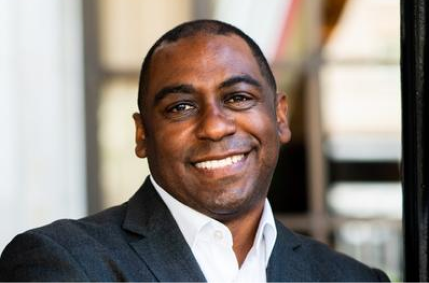 A professionally dressed man in a dark suit and white shirt, smiling confidently in an outdoor setting.