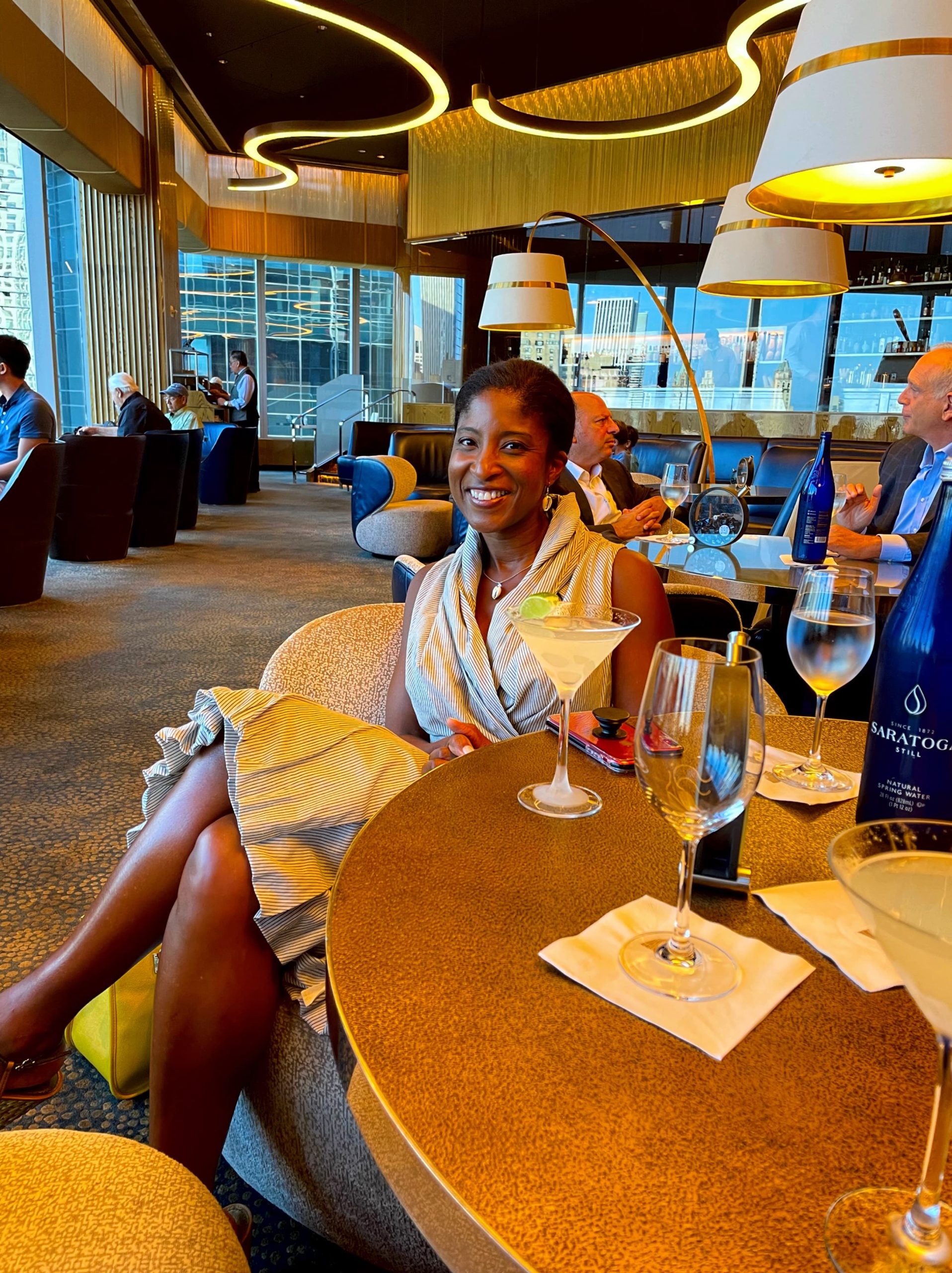 Smiling woman seated at a table in a stylish lounge with a cocktail and glasses of water, surrounded by a modern interior and city views.