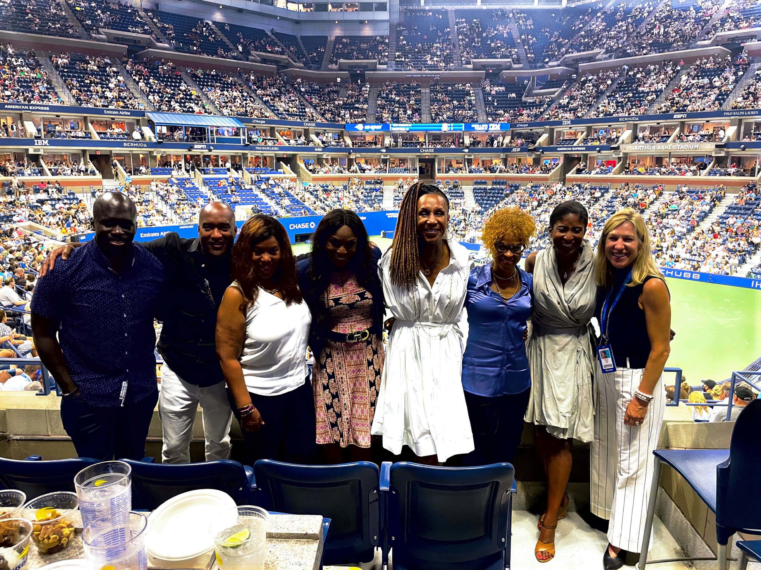Group of eight individuals smiling and posing in a stadium with a tennis court and a large crowd in the background.
