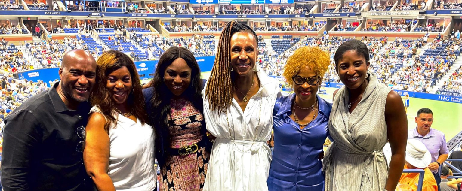 Group of six individuals smiling and posing in a stadium with a tennis court and a large crowd in the background.