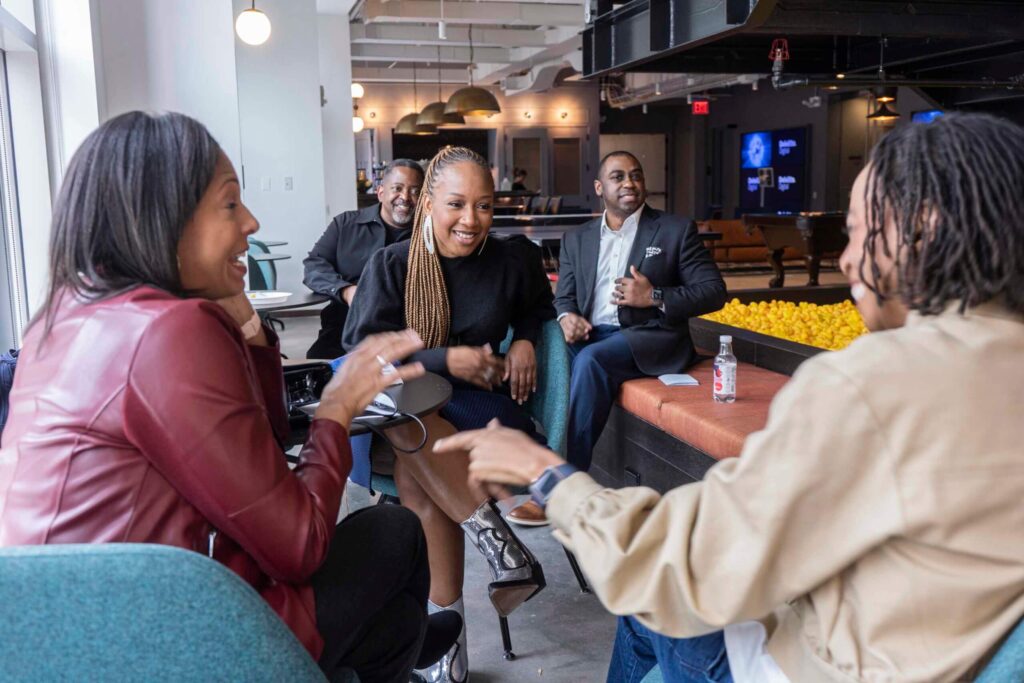 A group of five people laughing and talking in a modern lounge area with large windows and casual seating.