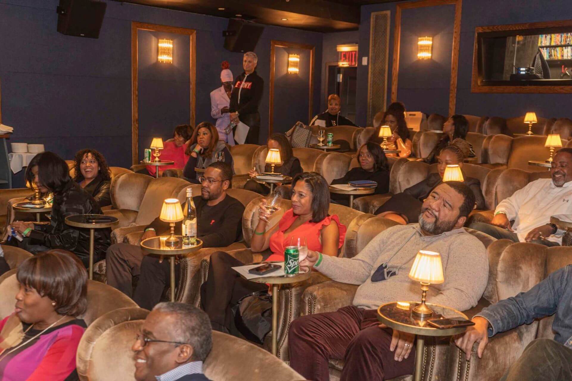 An attentive audience fills plush velvet chairs in a lounge, each seat accompanied by a small lamp, as people watch the event and sip beverages.