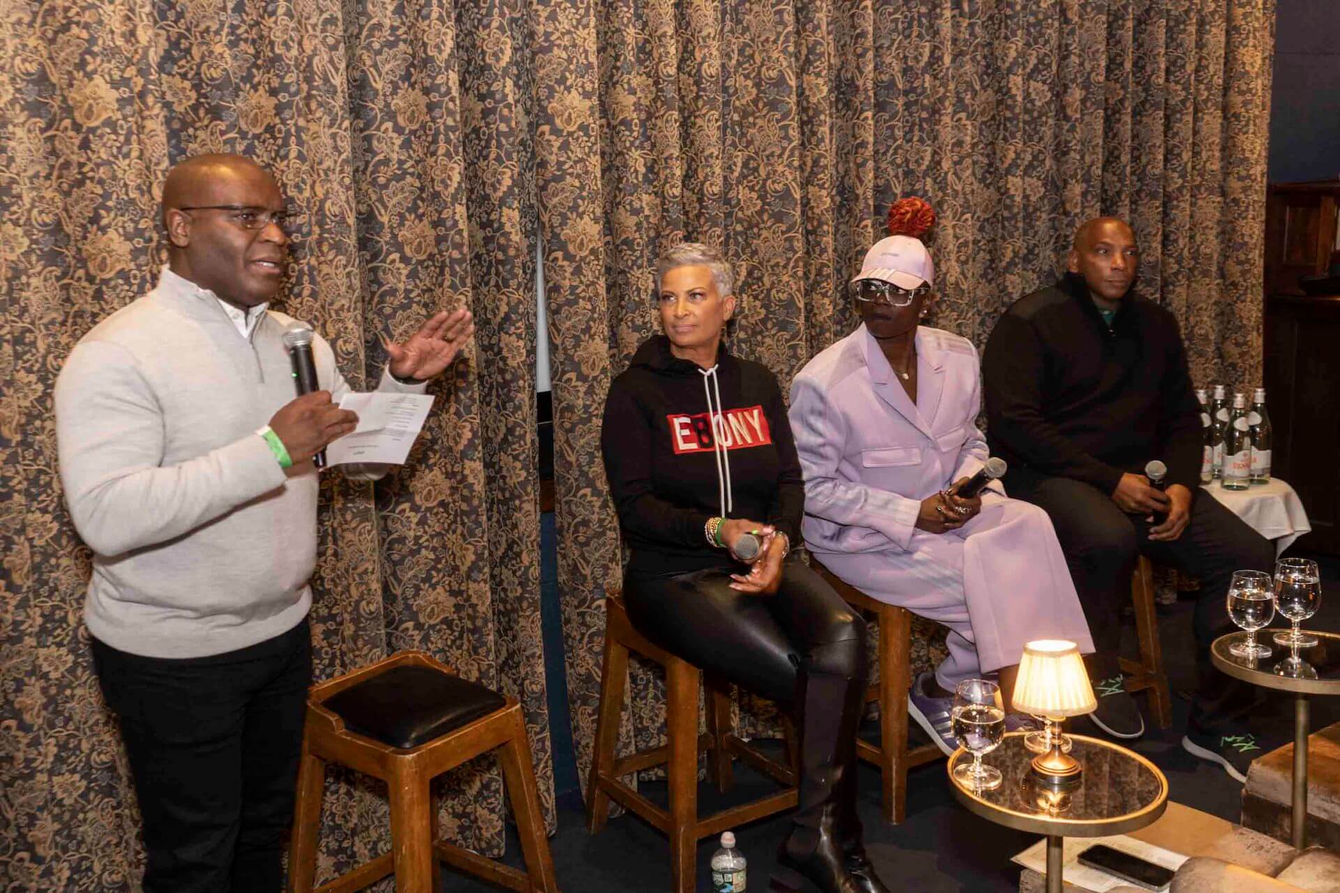 A man in a light-gray pullover speaks into a microphone, standing beside three seated panelists—one in an “Ebony” hoodie, another in a pastel suit and cap, and a third in a dark sweater—against a patterned curtain backdrop.