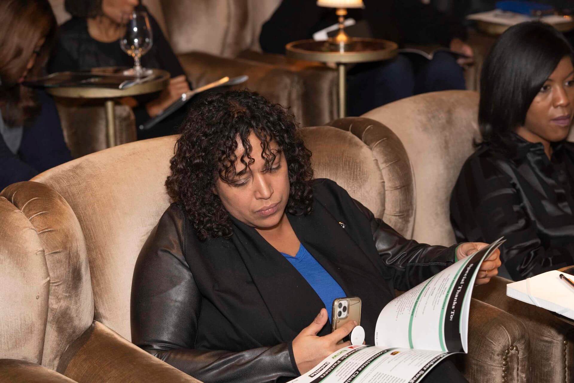 A curly-haired woman in a black jacket and blue shirt studies a booklet in her seat, her phone resting in her hand.