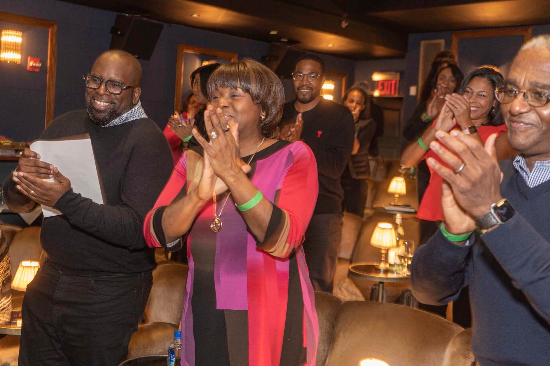 A standing audience claps enthusiastically, with a woman in a bright, color-blocked top leading the applause.