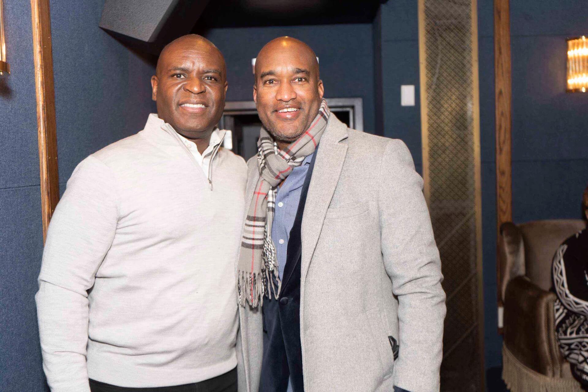 Two men wearing light-colored pullovers and blazers smile and pose together in a dimly lit lounge setting.