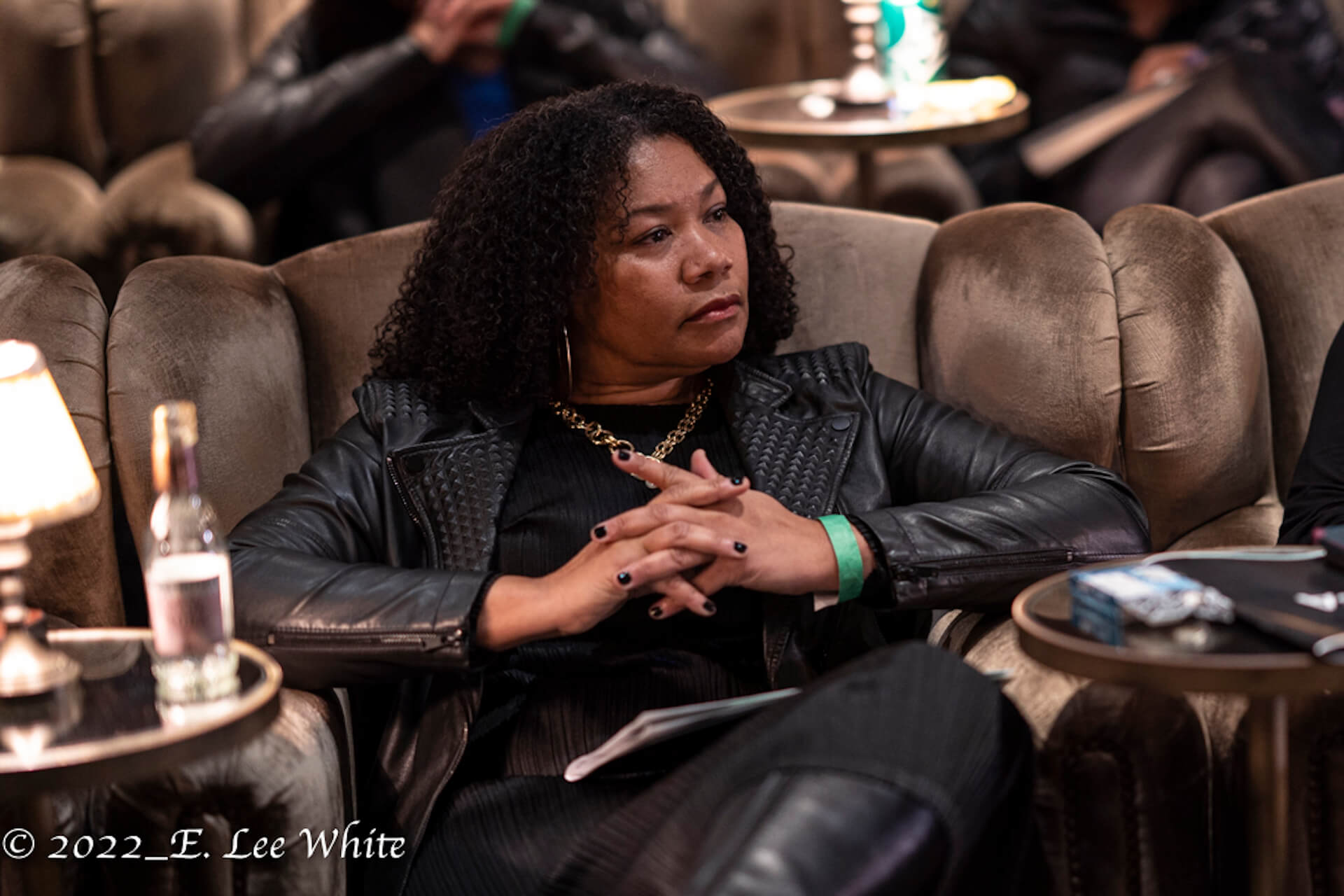 A woman in a black leather jacket sits in a plush chair, hands folded, attentively observing an event.