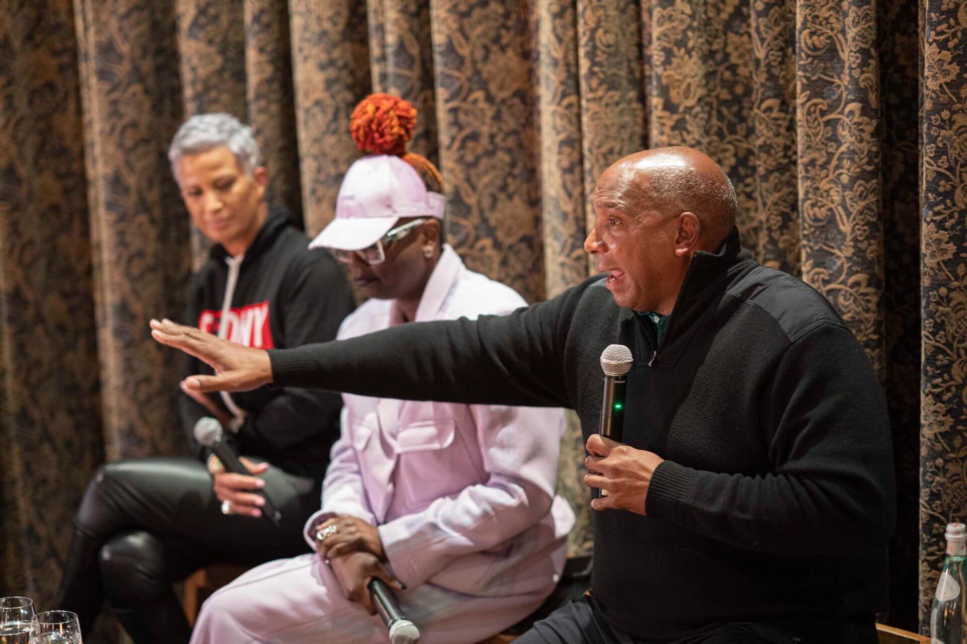 Three panelists seated in front of patterned curtains, with a man in a black sweater holding a microphone and extending his arm as he speaks. One panelist wears a pink cap and suit, and another wears a black hoodie.