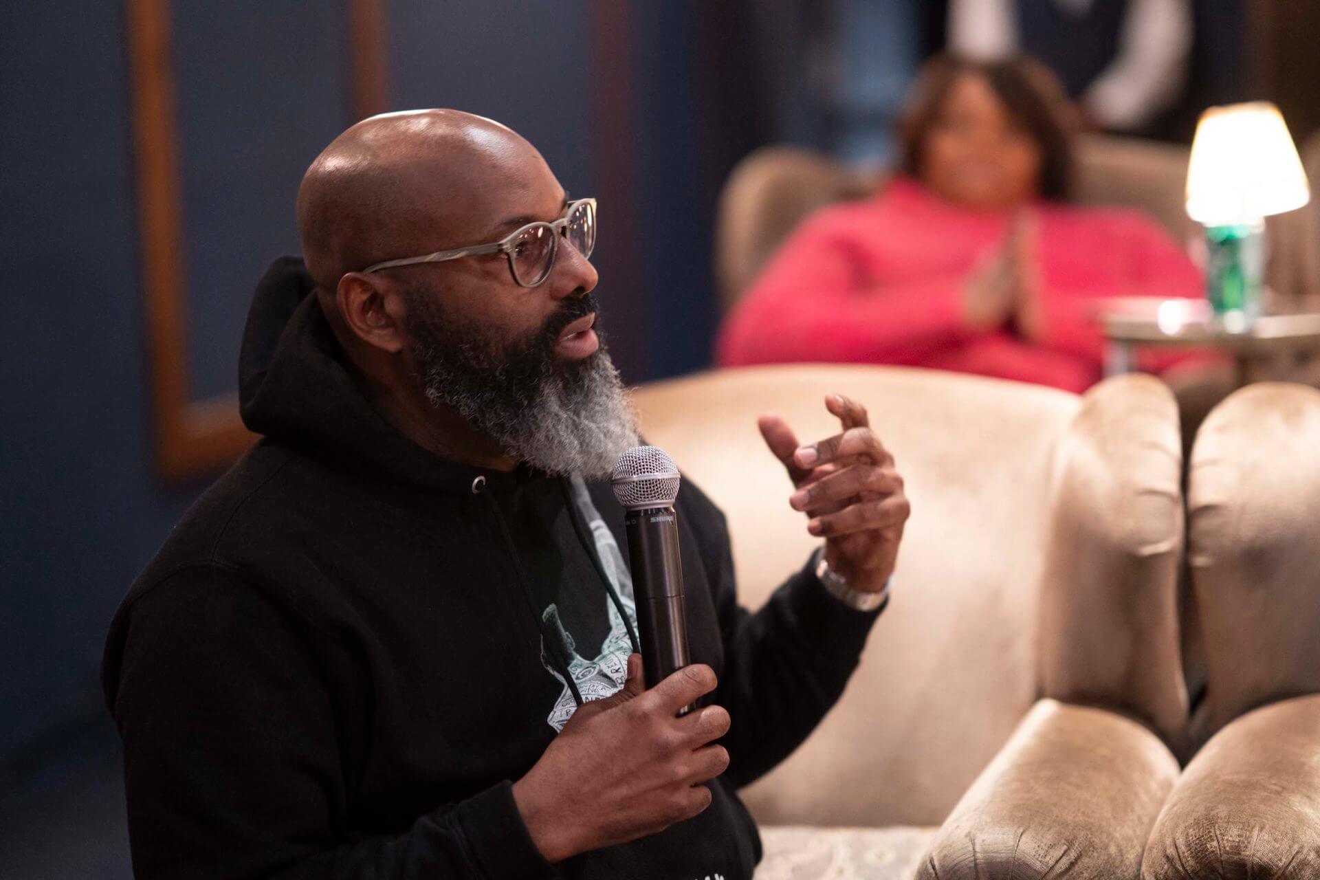 A bearded man in a black hoodie holds a microphone while seated in a velvet chair, gesturing as he addresses the gathering.