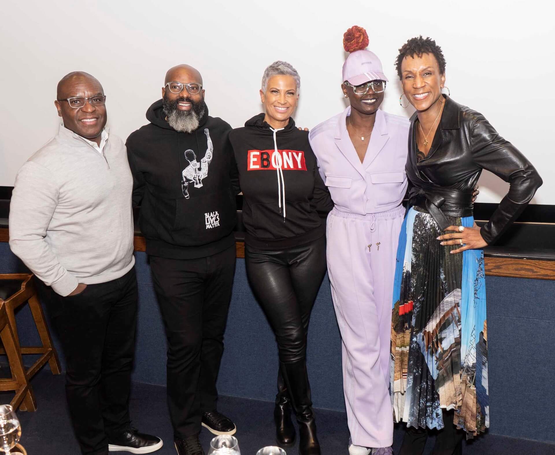 Five people stand together, smiling for the camera. One wears a light-gray pullover, another a black “Black Lives Matter” hoodie, another an “Ebony” hoodie, one in a lilac jumpsuit and cap, and one in a black leather top with a patterned skirt.