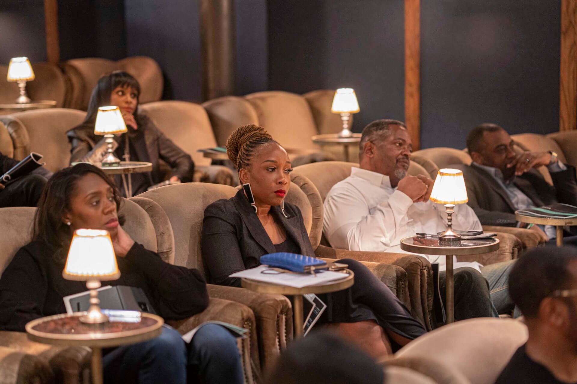 A group of attendees seated in plush lounge chairs, with a woman in a black blazer and braided updo in the foreground, all watching intently under soft lamp lighting.