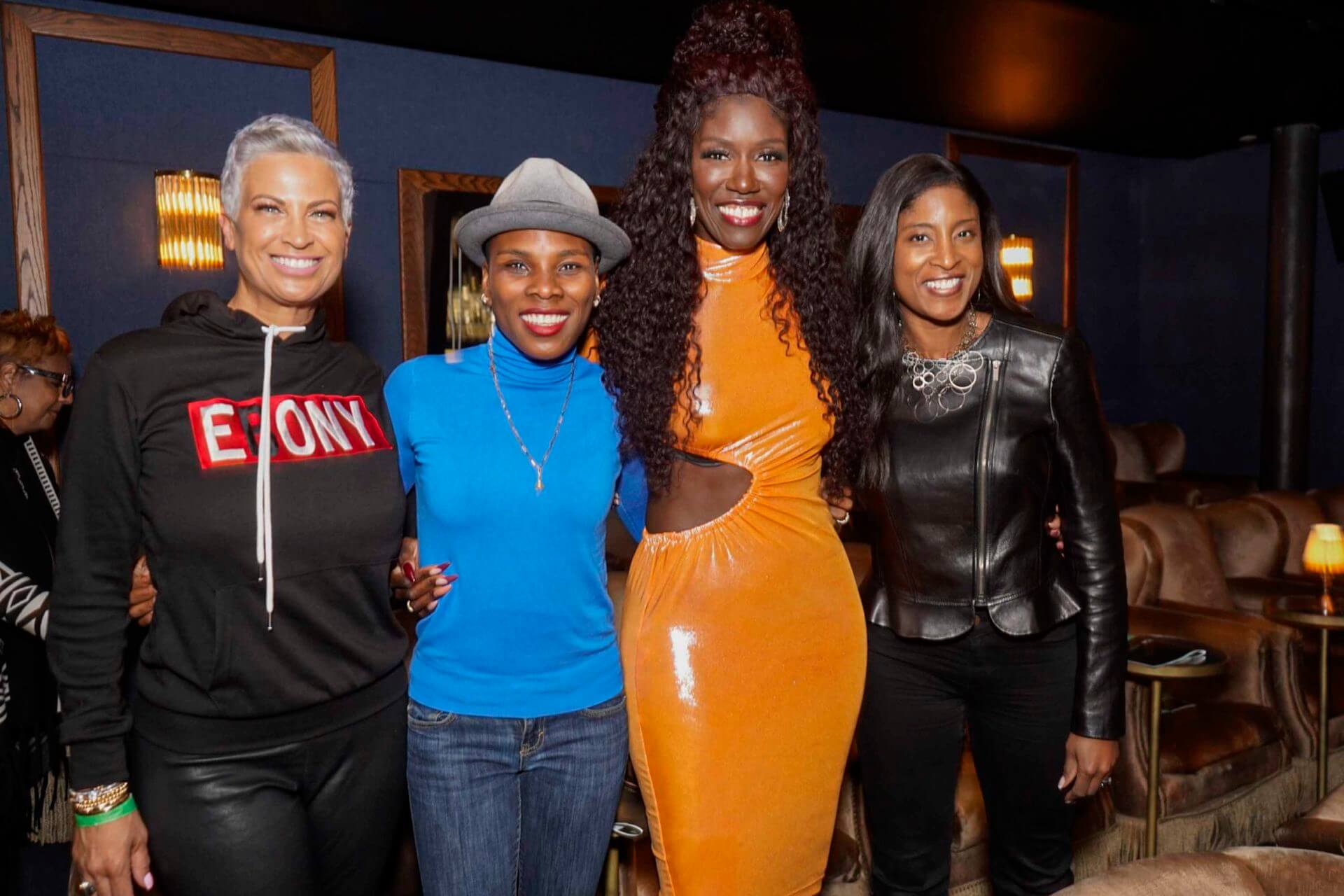 Four women posing and smiling; one wears an “Ebony” hoodie, another a bright orange cutout dress, another a hat with a blue turtleneck, and the fourth a black leather jacket.