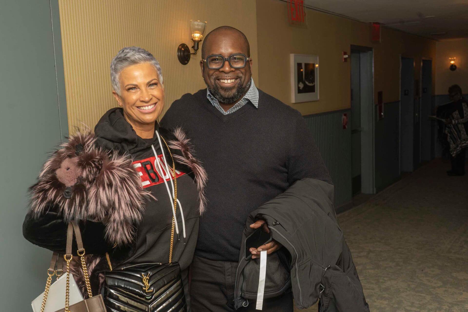 A smiling woman in a fur-trimmed hoodie stands beside a man in glasses and a dark sweater, posing together in a hallway.