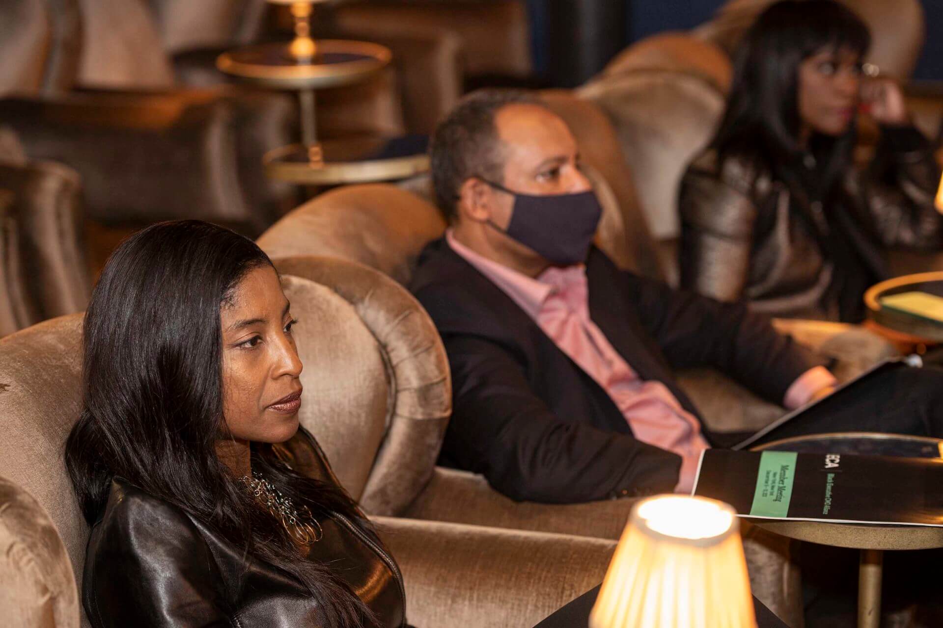 A woman in a leather jacket sits attentively in a velvet chair while a man in a mask leans back beside her in the softly lit lounge.