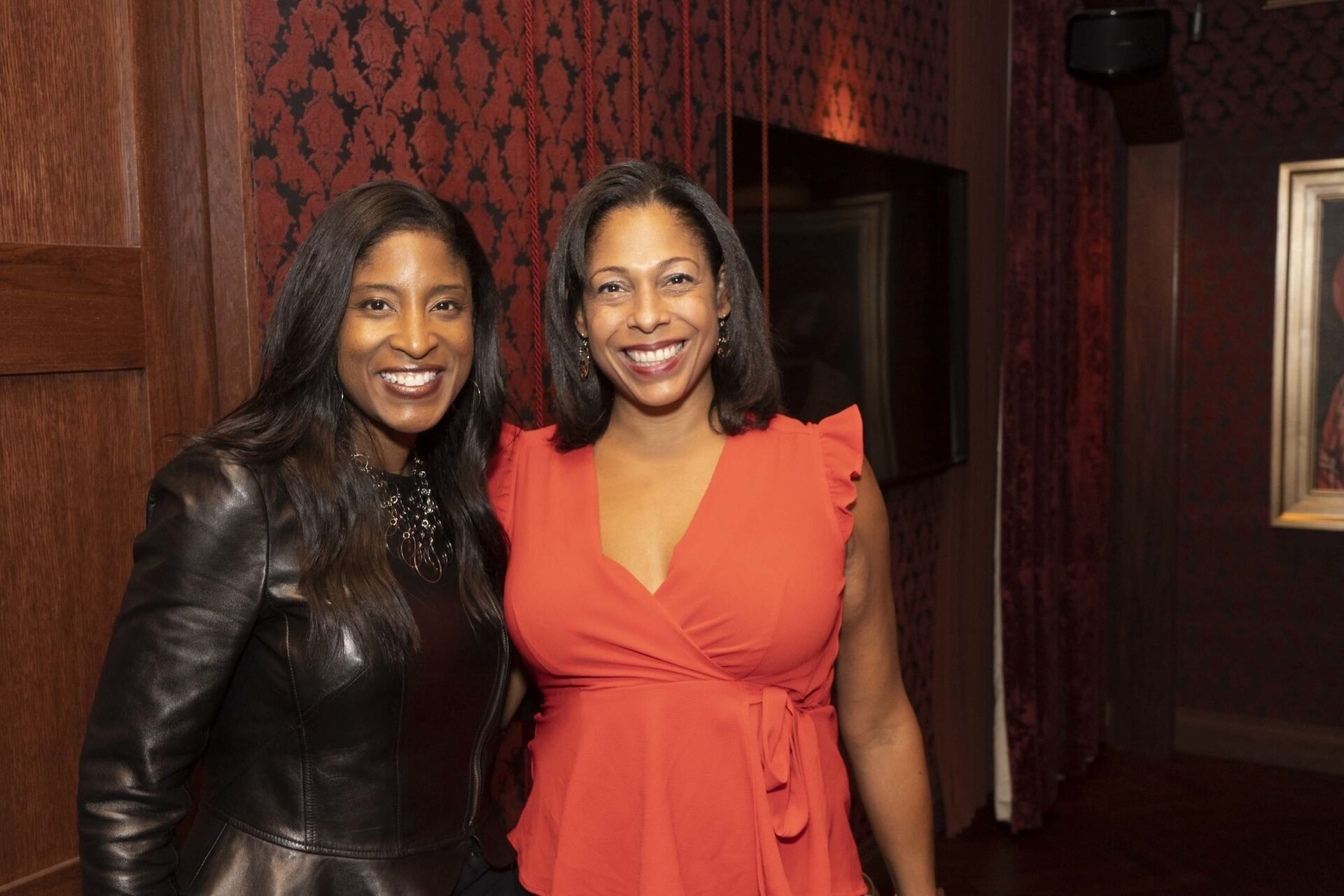Two women smiling and posing together in a warmly lit room, one wearing a black leather jacket and the other in a red blouse with ruffled sleeves