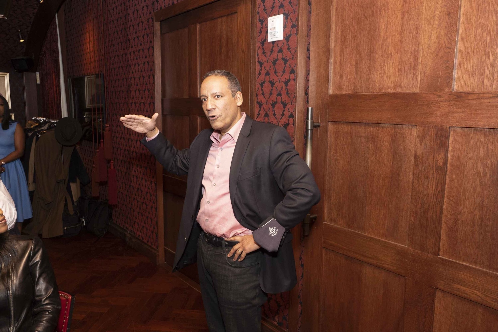 Man speaking and gesturing with his hand in a room with wooden doors and red patterned walls, with a clothing rack in the background.