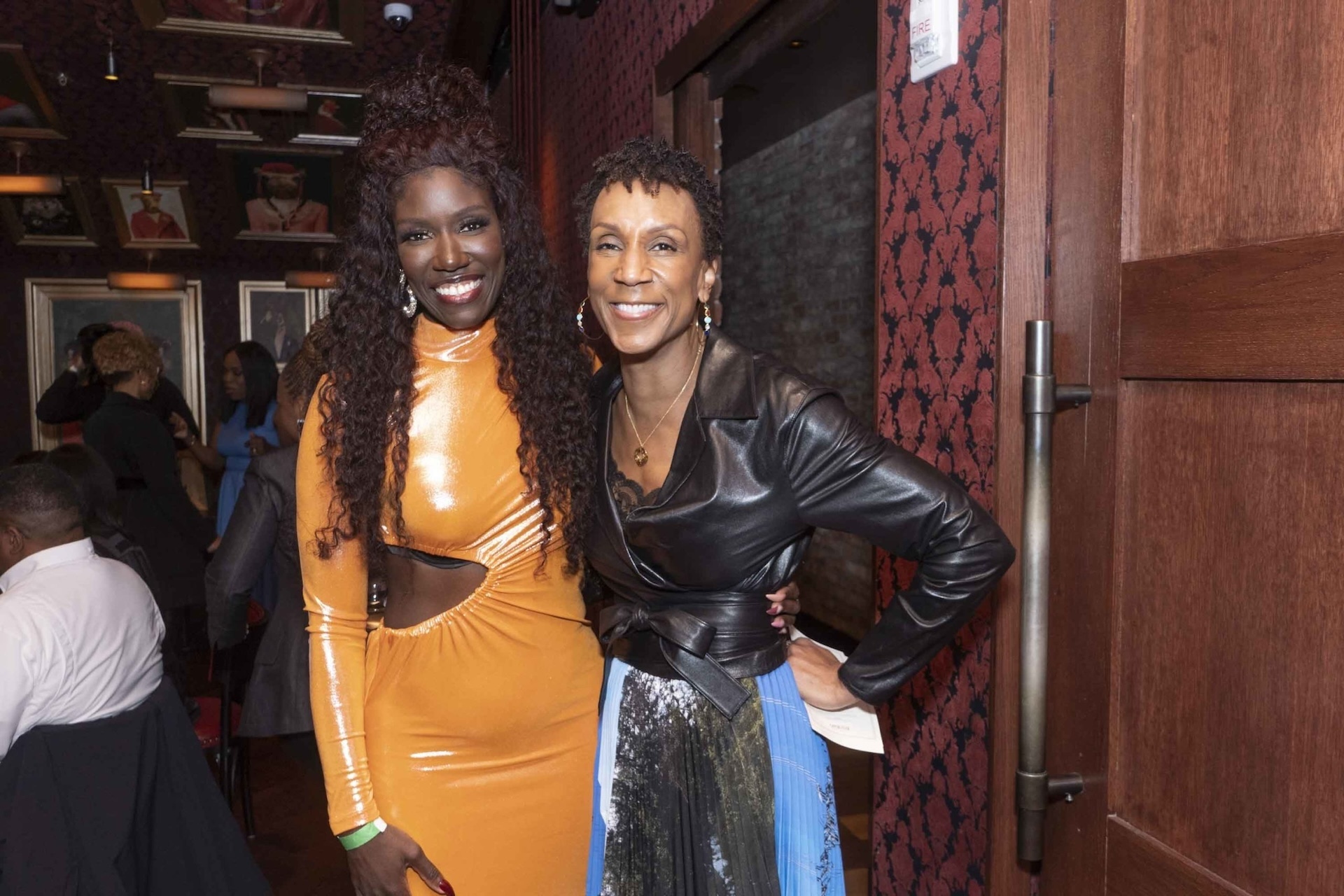 Two women smiling and posing, one wearing a shiny orange dress and the other in a leather jacket and patterned skirt, in a warmly lit room.
