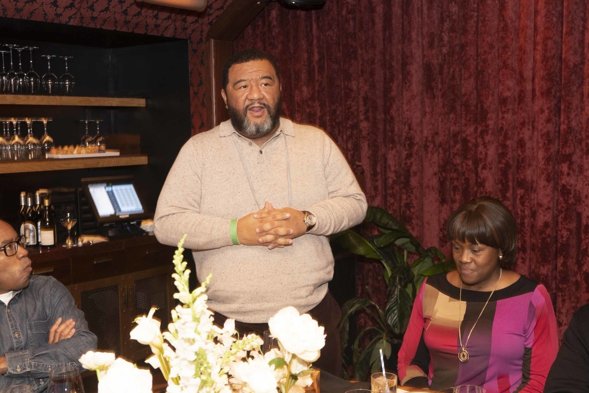 Man standing and speaking in front of a table with white flowers, with other guests listening, in a warmly lit room with red velvet curtains.