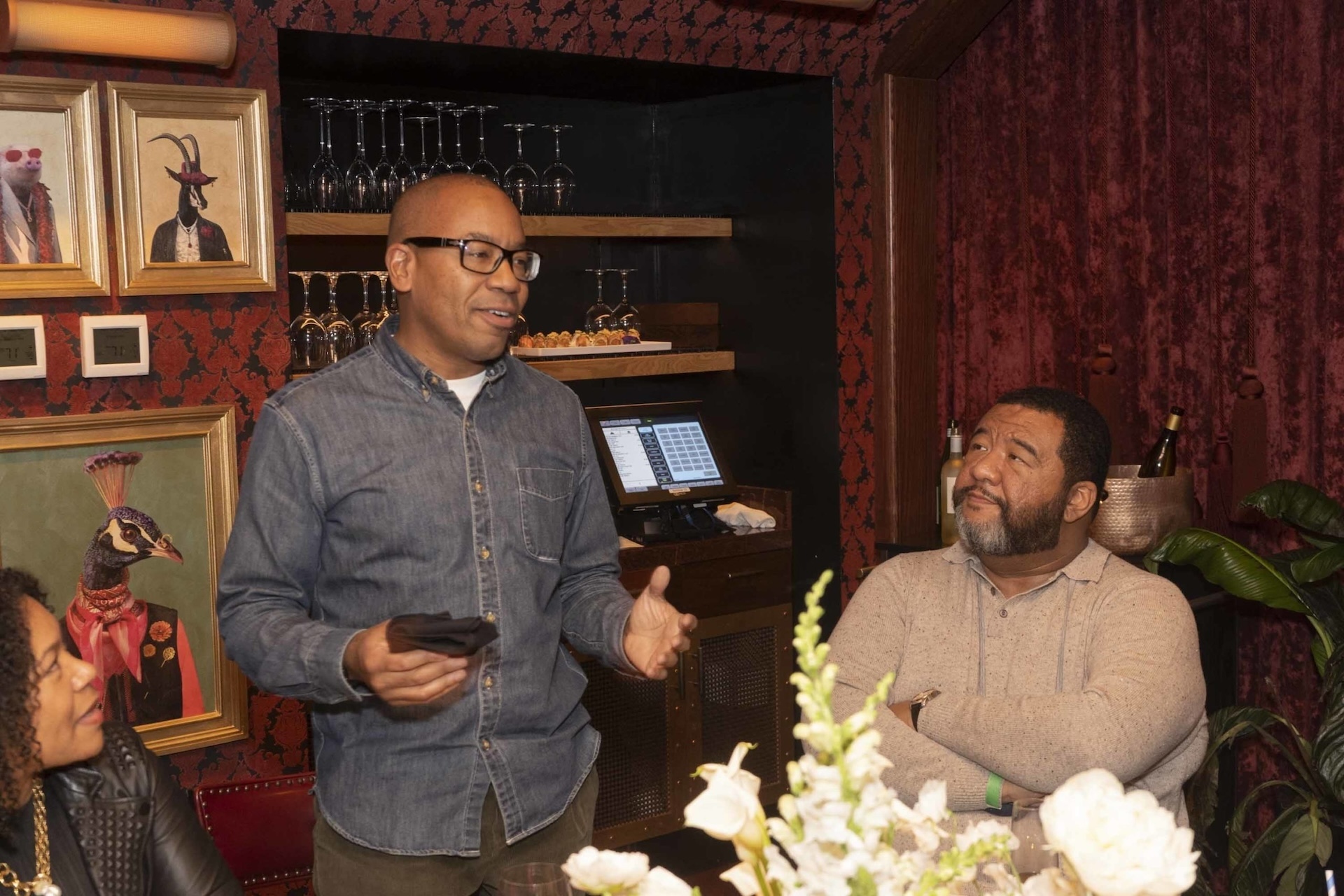 Man speaking while gesturing with hands in a warmly lit room with framed artwork and wine glasses displayed on shelves in the background
