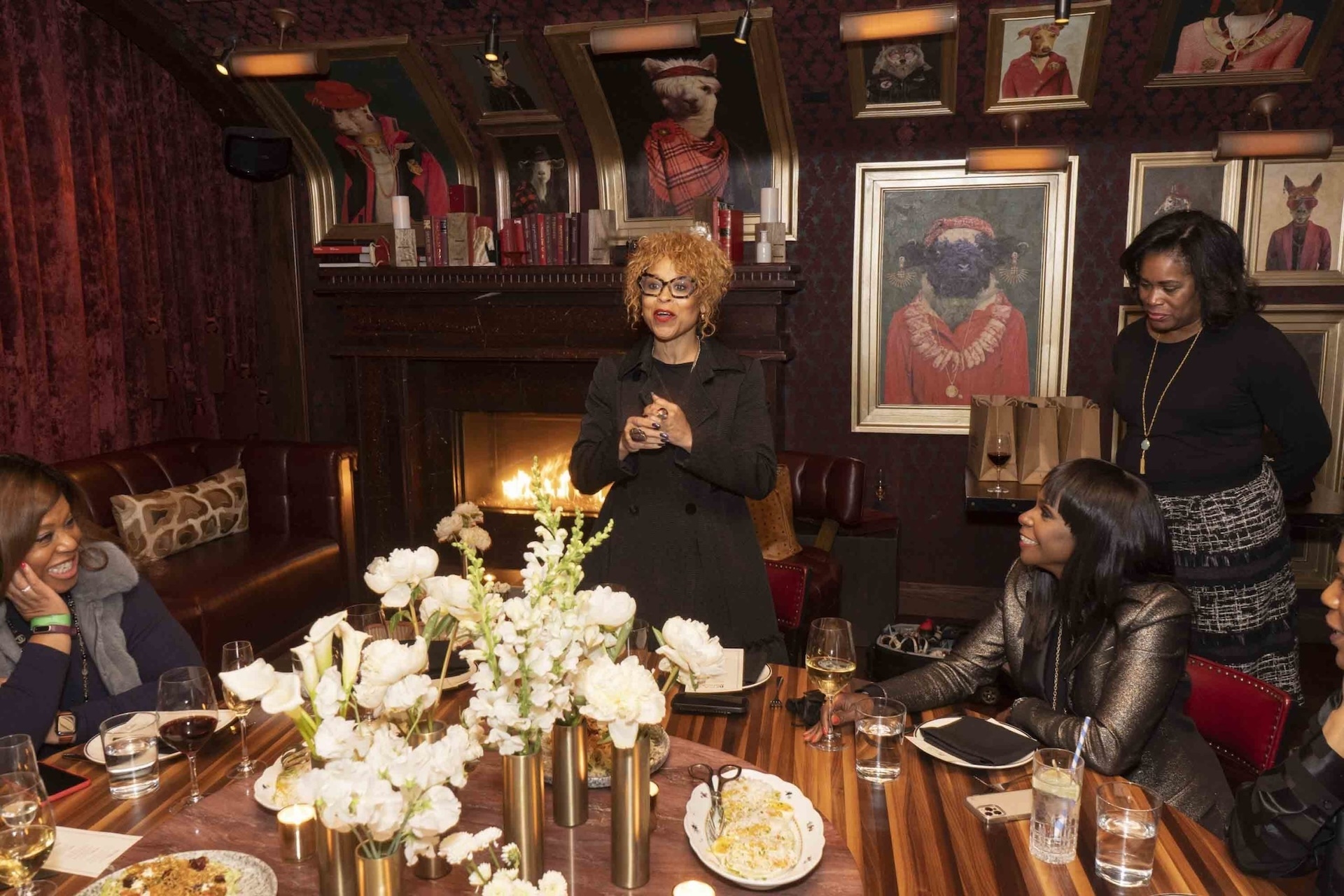 Woman speaking at a table with white floral centerpiece, fireplace, and framed artwork in the background in a cozy room