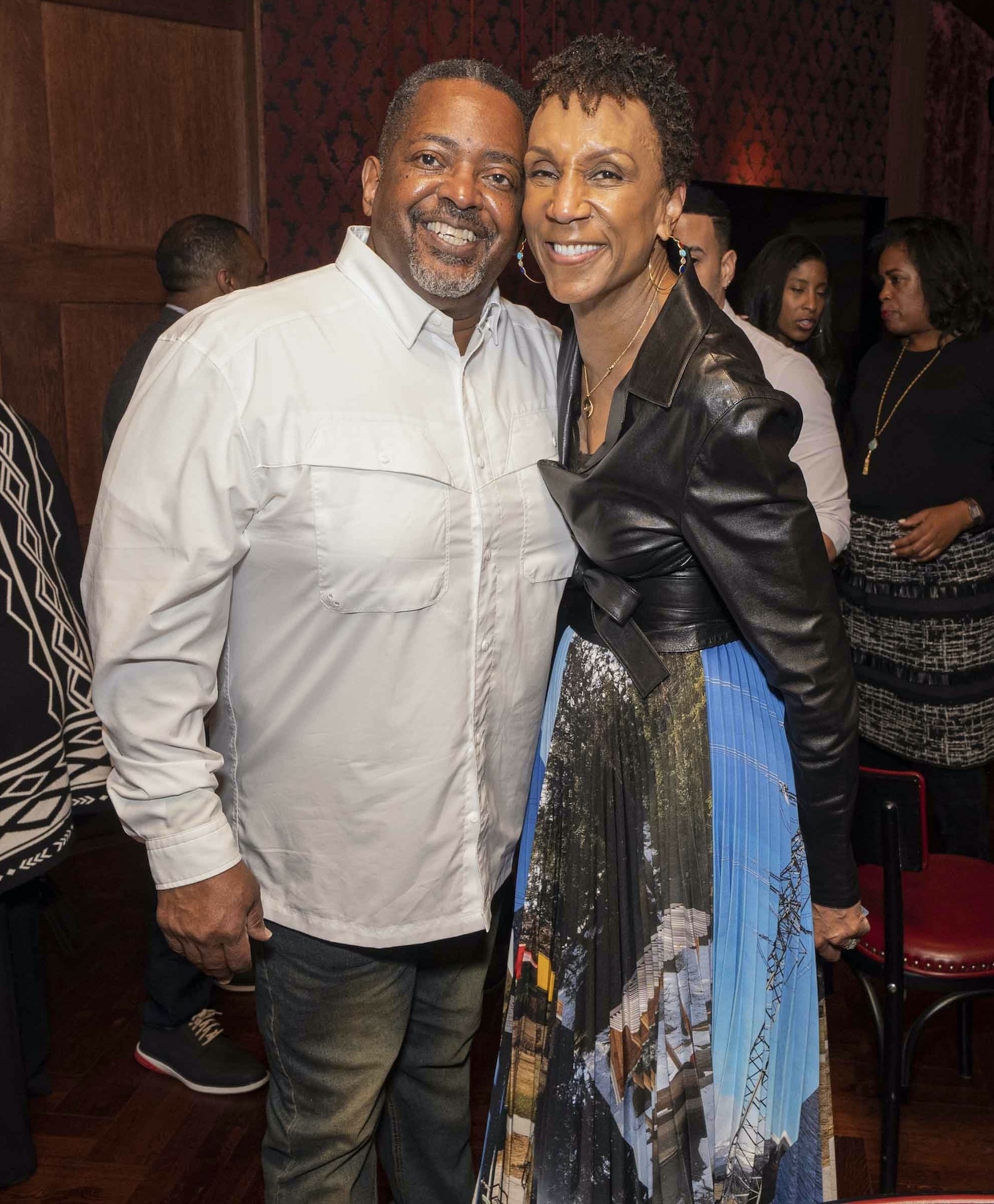 Man and woman smiling and posing together in a warmly lit room, the man in a white shirt and jeans and the woman in a leather jacket and colorful skirt.