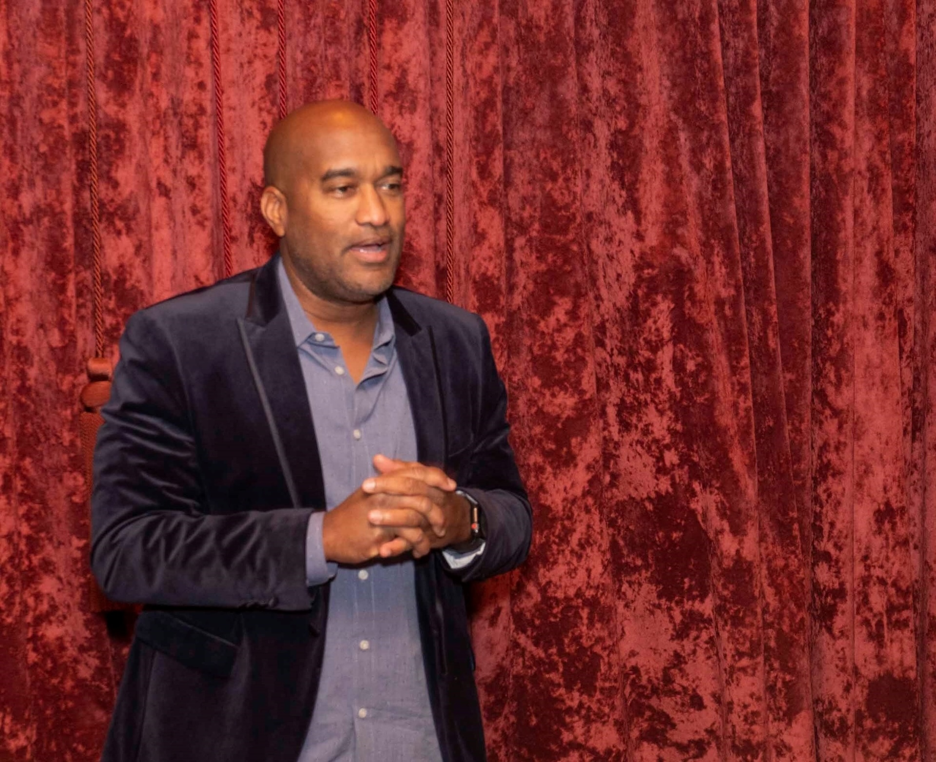 Man speaking in front of red velvet curtains, gesturing with his hands, in a warmly lit room.