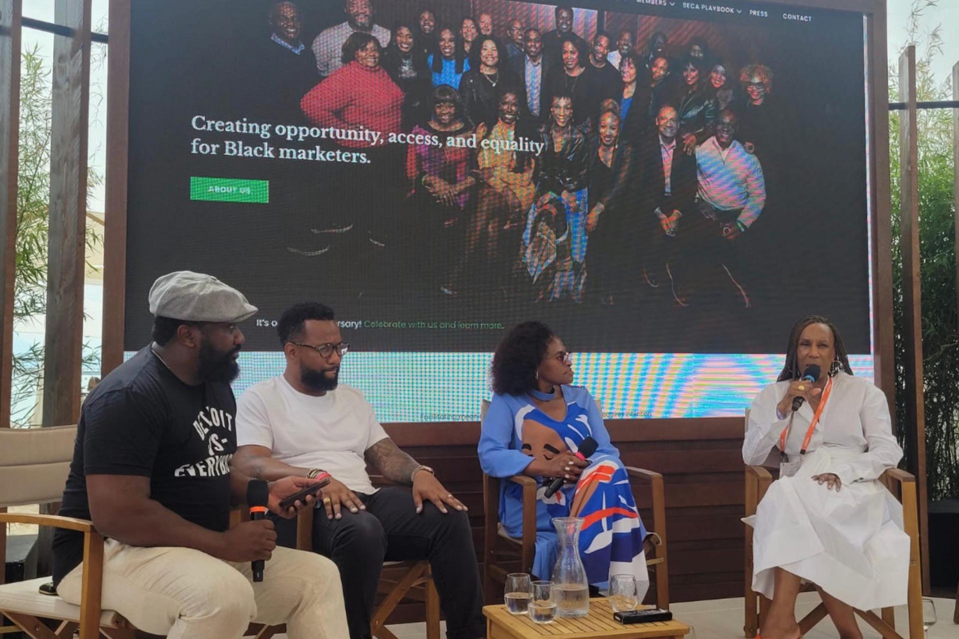 Four panelists are seated onstage in front of a large screen that reads “Creating opportunity, access, and equality for Black marketers,” as one woman in a white dress speaks into a microphone.