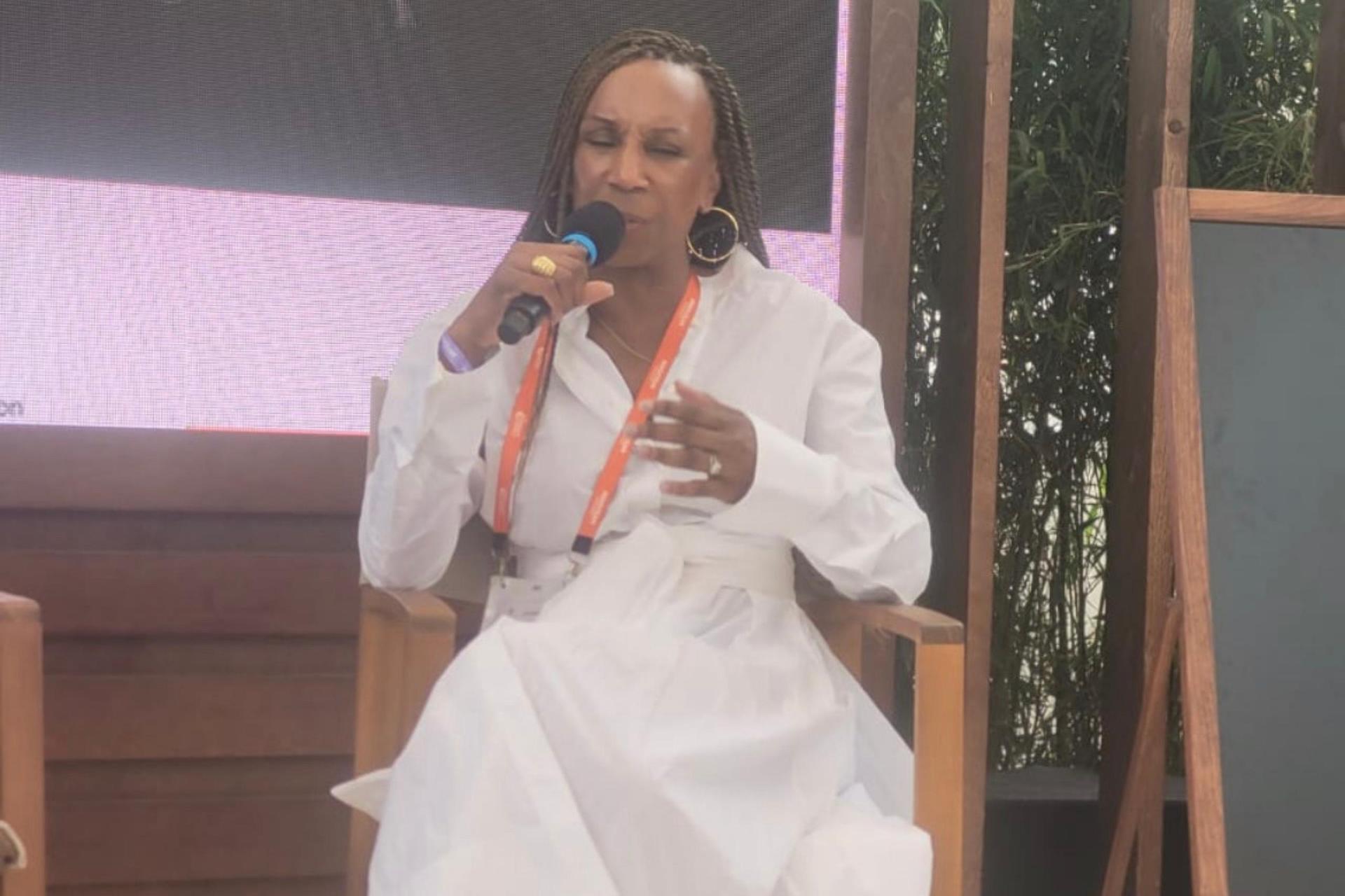 A close-up of the same woman with braided hair in a white belted dress, holding a microphone and speaking during the discussion.