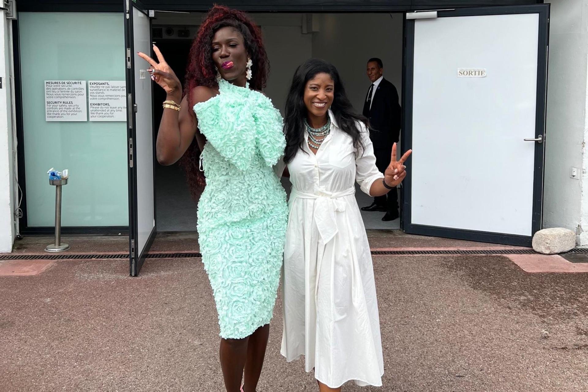 Two women pose outside a venue, one in the mint-green floral dress making a playful peace sign, and the other in a white belted dress also flashing a peace sign.