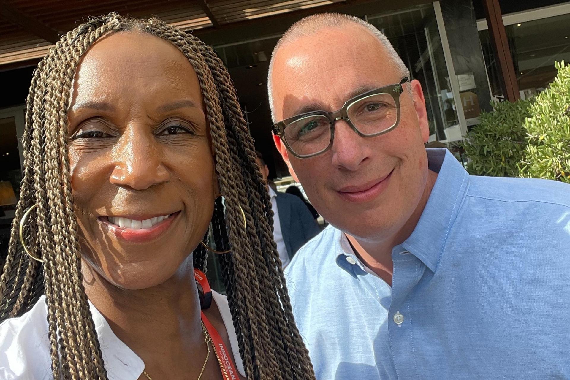A close-up selfie of a woman with braided hair in a white shirt smiling next to a man in glasses and a light-blue button-down, both looking at the camera.
