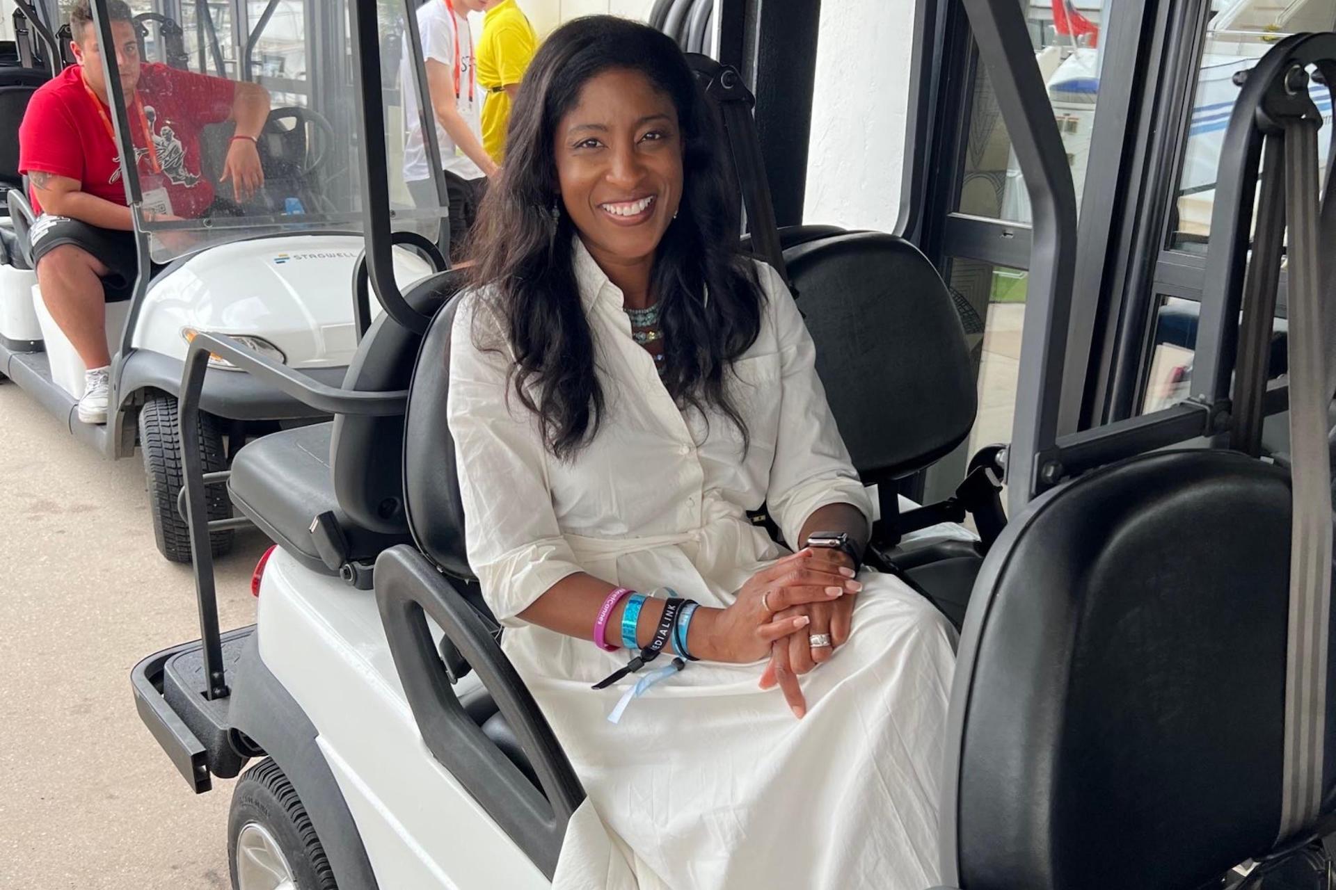 A smiling woman wearing a white dress sits in a golf cart, hands folded, adorned with colorful wristbands, with others nearby in the background.