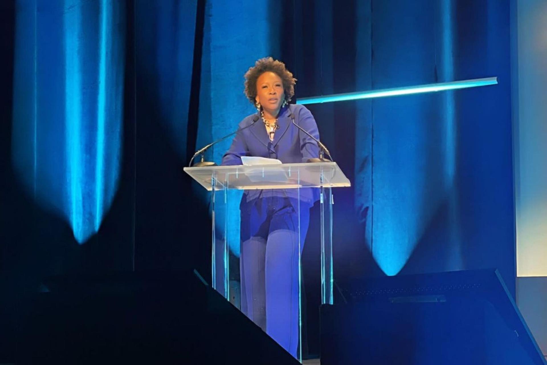 A woman in a bright-blue suit speaks at a clear podium on a stage illuminated with blue lights.
