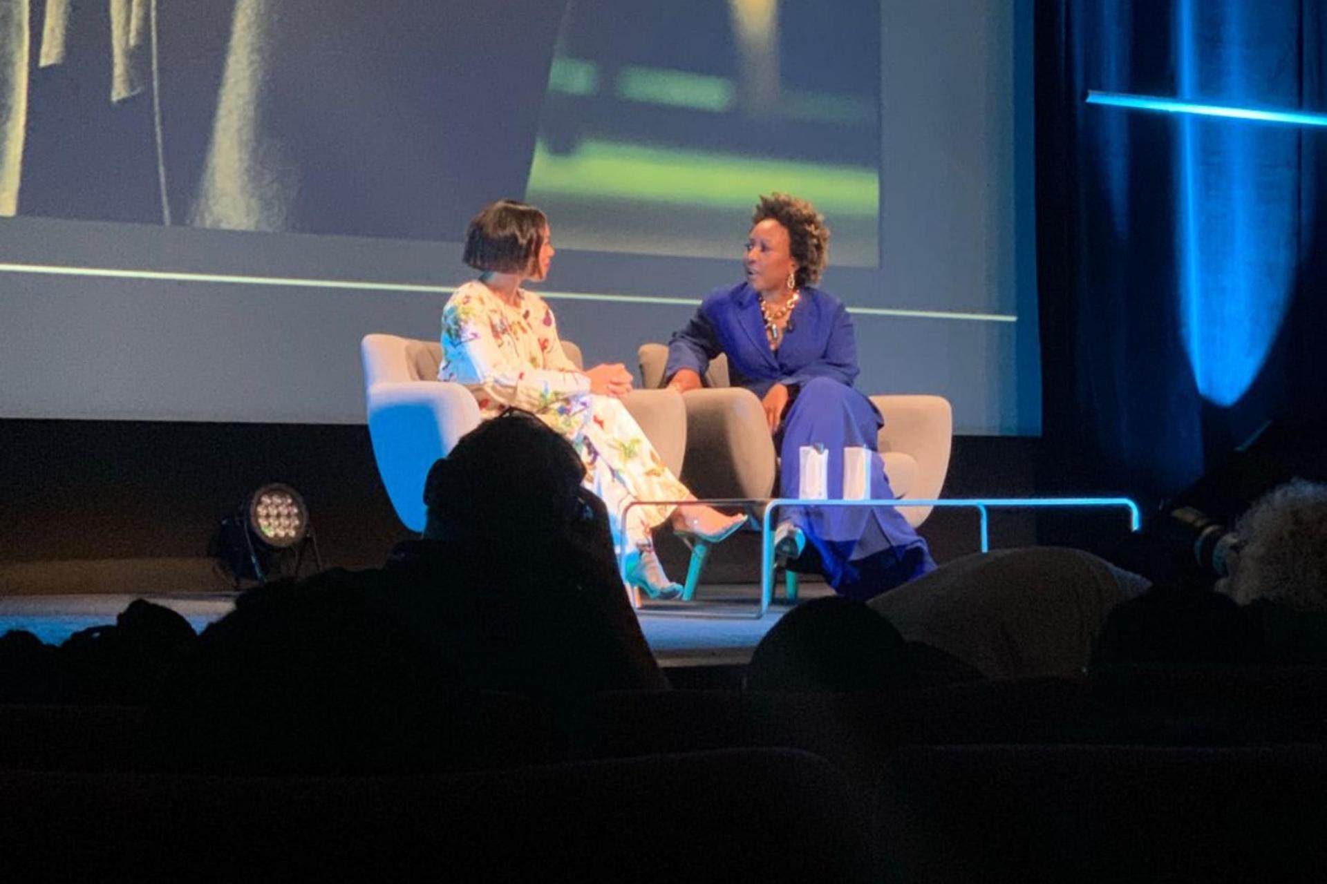 Two women seated on stage, one in a floral jumpsuit and the other in a blue suit, engaged in conversation.