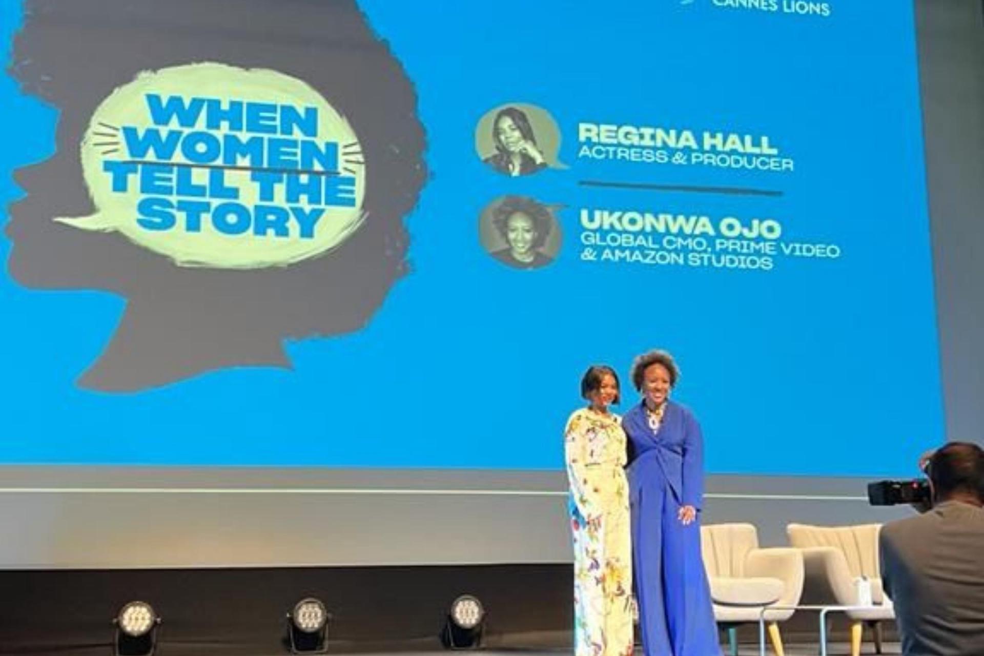 Two women stand on a stage under a large blue screen that reads “When Women Tell The Story,” displaying the names Regina Hall (actress & producer) and Ukonwa Ojo (Global CMO, Prime Video & Amazon Studios).