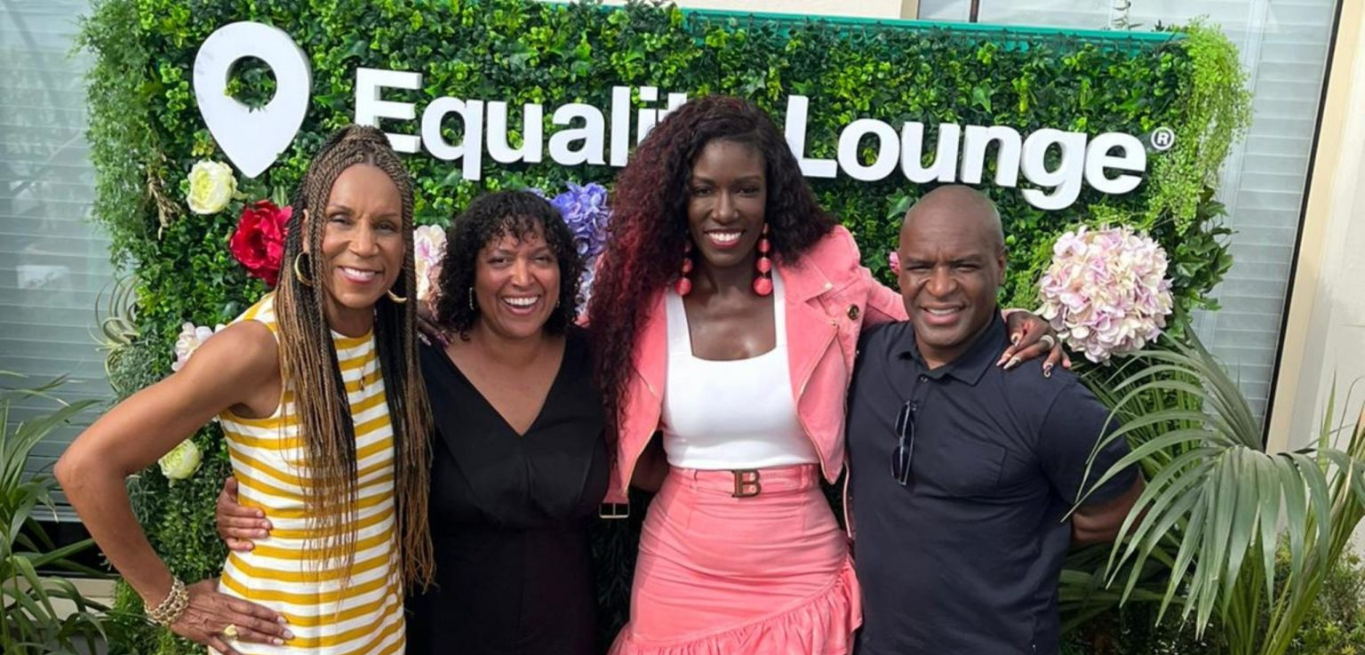 Four people pose together in front of a green floral wall with the words "Equality Lounge®" displayed. They are smiling and dressed in a mix of casual and stylish attire, with one person wearing a bright pink outfit. The group appears to be at an outdoor networking or professional event, with greenery and decorative flowers in the background.