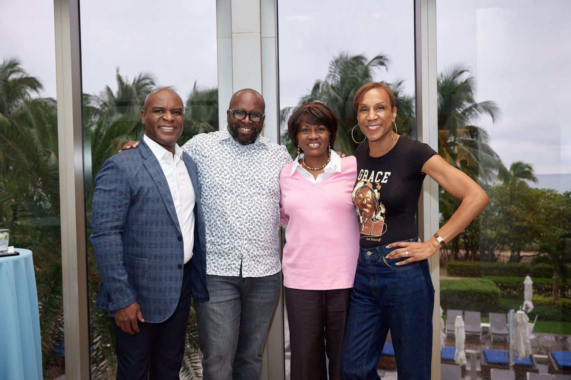 Four people standing side by side near tall windows overlooking palm trees; one in a blue blazer, another in a patterned shirt, another in a pink sweater, and the last wearing a “Grace” T-shirt.