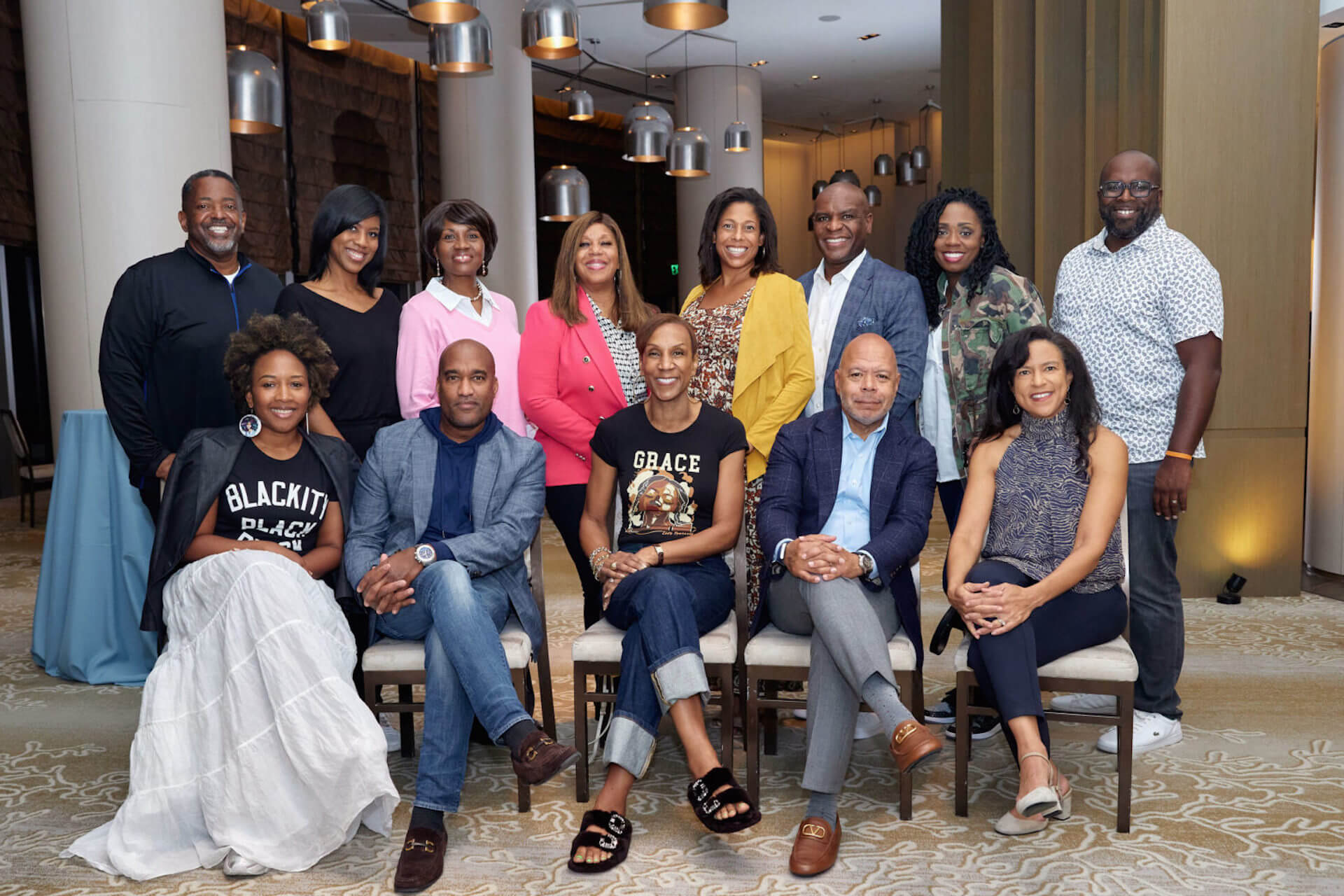 A group of twelve people posing in a hotel lobby, some seated, some standing. The front row features one person in a “Blackity Black Black” T-shirt and another in a “Grace” T-shirt.