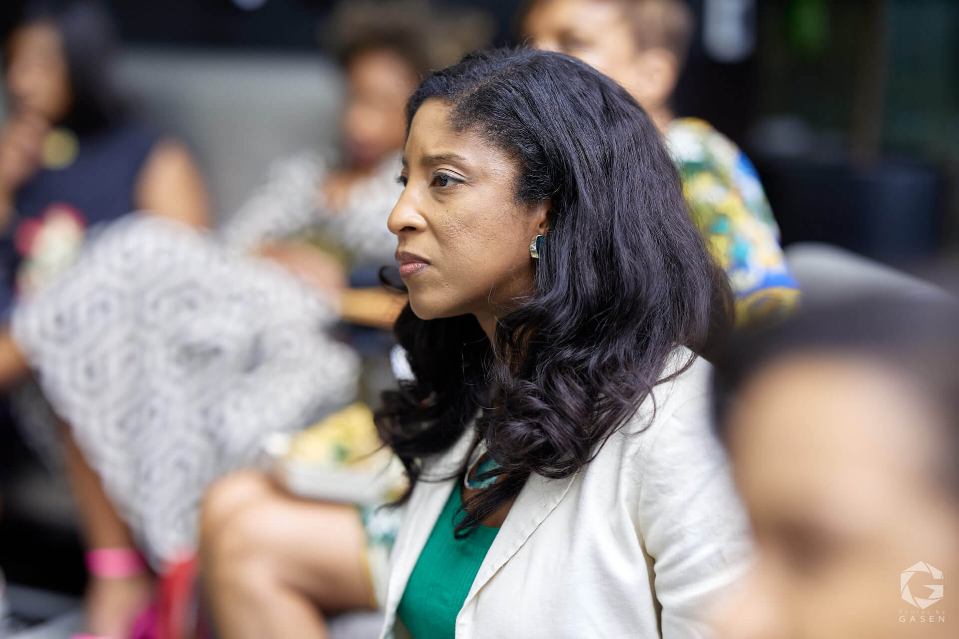 A woman with long dark hair, dressed in a light-colored blazer, watches the discussion attentively.