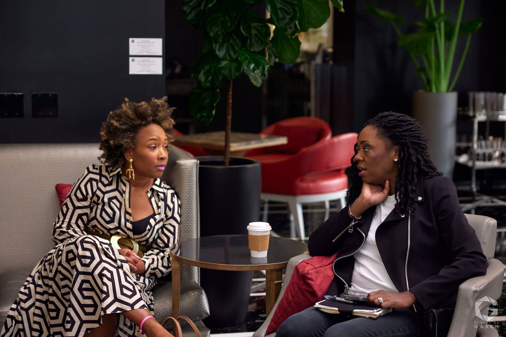 Two women seated in a hotel lounge, deep in conversation, with one wearing a black-and-white geometric print dress and the other in a black jacket, a coffee cup on the table between them.