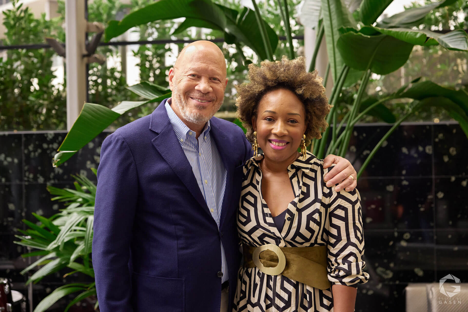A man in a navy blazer poses beside a woman in a black-and-white patterned dress with a wide belt, both smiling against a backdrop of tall tropical plants.