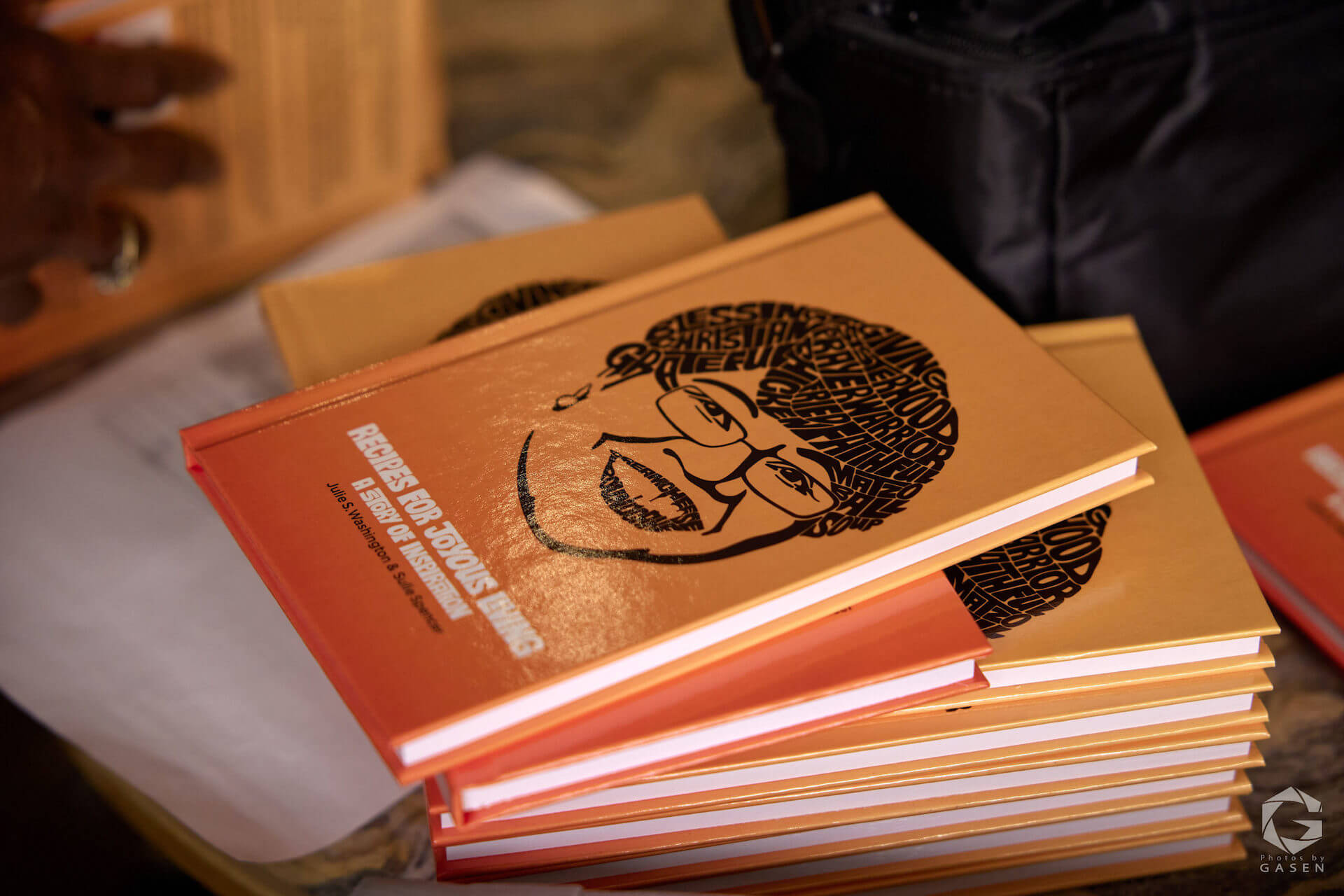 A close-up of several orange hardcover books titled “Recipes for Joyous Living: A Story of Inspiration,” featuring a stylized illustration of a woman’s face on the cover.