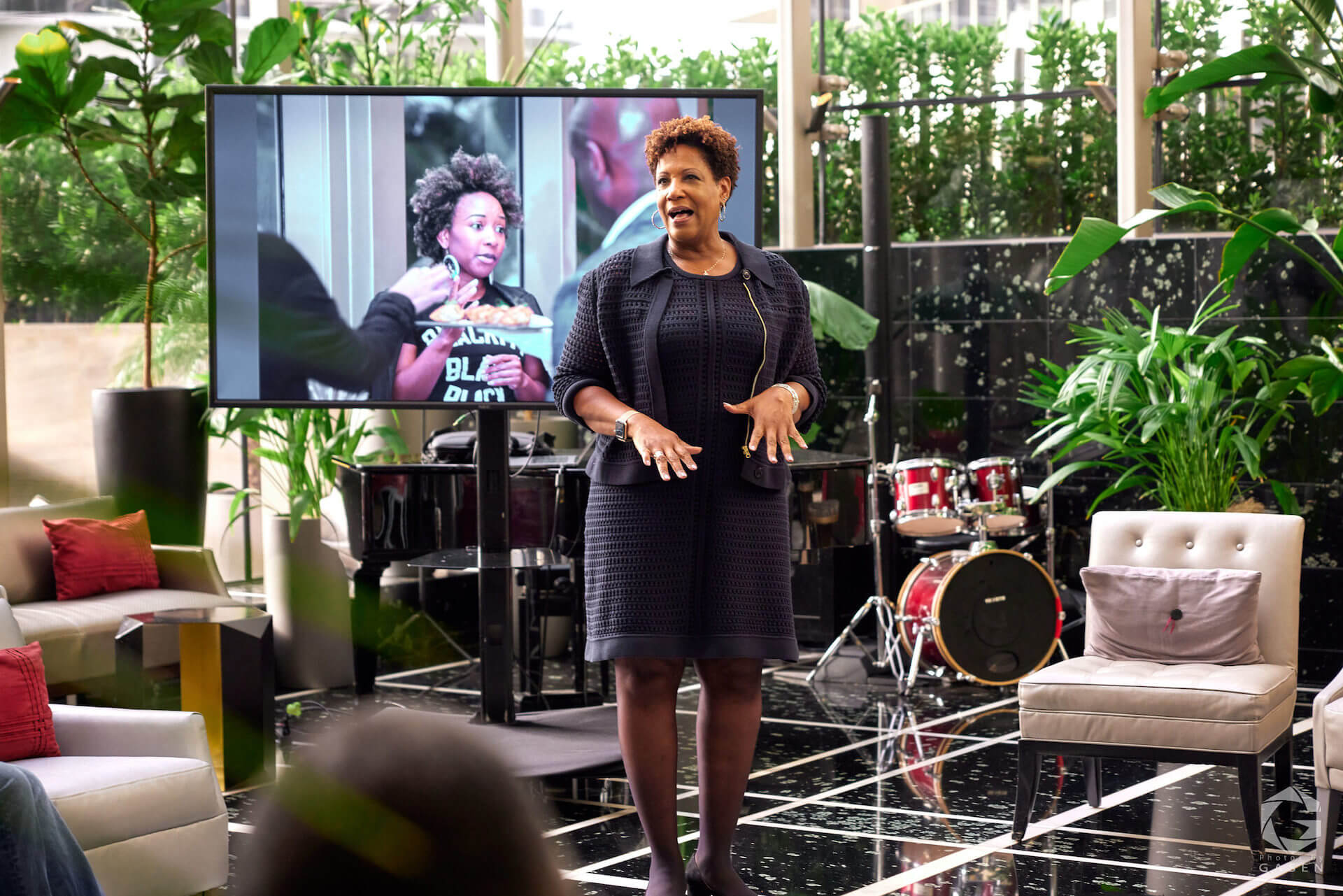 A woman in a textured black dress speaks in front of a large screen displaying a photo of another attendee wearing a “Blackity Black Black” T-shirt. She stands in a stylish lounge with a piano and drum set behind her.