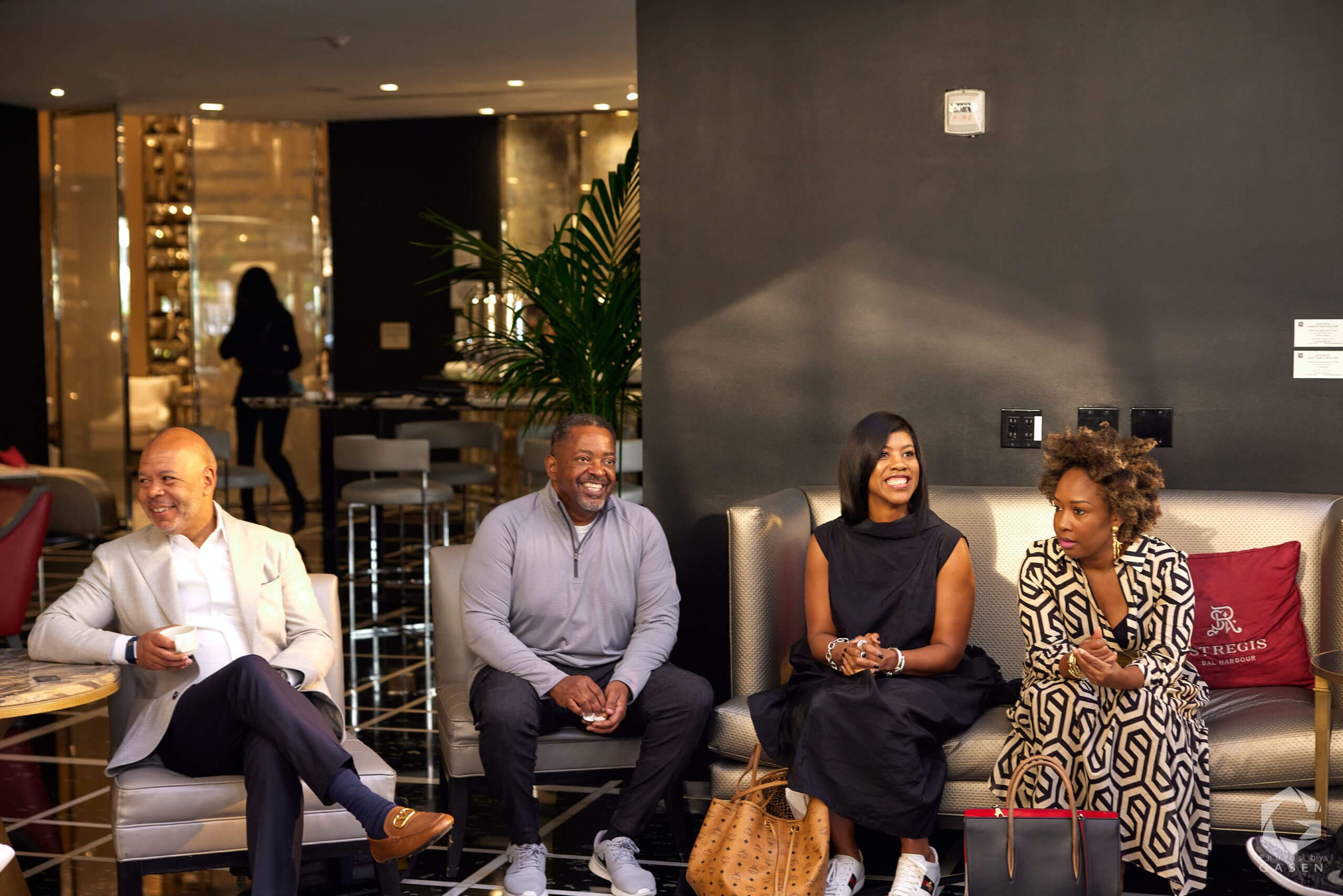 Four people seated in a row: a man in a light-gray blazer, a man in a gray pullover, and two women—one in black, another in a geometric-print dress—all laughing and conversing.